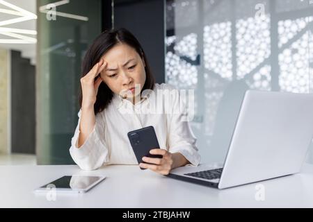 Femme triste bouleversée au lieu de travail tenant un smartphone, femme d'affaires déprimée naviguant sur les médias sociaux à l'intérieur du bureau, travailleuse asiatique utilisant un ordinateur portable au travail. Banque D'Images