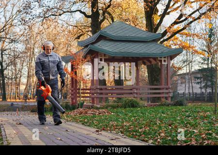 Homme d'entretien travaillant dur enlevant les feuilles jaunes du trottoir à l'extérieur. Vue d'angle bas du travailleur masculin focalisé dans l'âge utilisant le ventilateur de feuille dans le parc, avec gazebo sur le fond. Concept de travail saisonnier. Banque D'Images