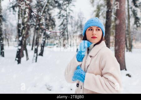 Gros plan portrait de jeune femme marchant sous la neige dans le parc d'hiver portant manteau de fourrure blanche, chapeau bleu tricoté et mitaines. Vêtements chauds et confortables Banque D'Images