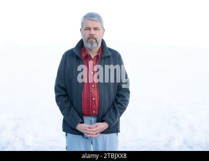 Homme, 56 ans, cheveux gris et barbe, debout dehors en hiver avec de la neige en arrière-plan, Banque D'Images