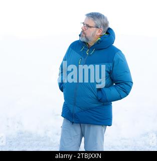 Homme, 56 ans, cheveux gris et barbe, debout dehors en hiver avec de la neige en arrière-plan, les mains en poche, portant des lunettes Banque D'Images