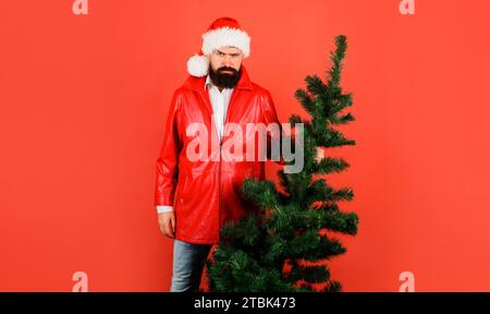 Homme sérieux dans le chapeau de Santa avec arbre de Noël. Joyeux Noël et bonne année. Homme barbu en manteau rouge avec sapin. Homme d'affaires se préparant pour New Banque D'Images