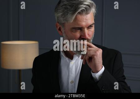 Bel homme barbu fumant le cigare près du mur gris foncé à l'intérieur Banque D'Images