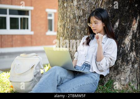 Une étudiante asiatique positive et réfléchie utilise son ordinateur portable sous l'arbre dans un parc du campus, faisant ses devoirs en ligne. Banque D'Images