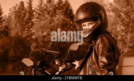 Moto girl chevauchant la moto de tourisme sur la route forestière. Image teintée dans la couleur Peach Fuzz de l'année 2024. Belle femme avec les cheveux rouges dans la veste en cuir Banque D'Images