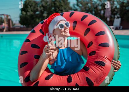 Chapeau de père Noël piscine femme. Une femme heureuse dans un bikini bleu, un chapeau de Père Noël rouge et blanc et des lunettes de soleil pose près de la piscine avec une coupe de champagne debout Banque D'Images