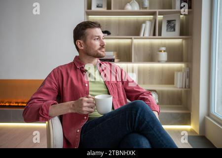 Homme positif calme assis dans un appartement moderne avec une tasse de thé, pensant, se reposant, regardant la fenêtre Banque D'Images