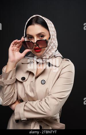 Élégante dame caucasienne regardant sur des lunettes de soleil à la caméra à l'intérieur. Portrait de femme à la mode sérieuse portant trench coat et foulard regardant droit, isolé sur noir. Mode, concept de style. Banque D'Images