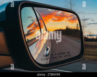 Coucher de soleil reflété dans le rétroviseur latéral du véhicule sur la route bordée d'arbres. Banque D'Images