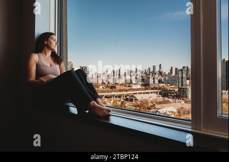 Femme assise sur le rebord d'une grande fenêtre regardant la grande ville. Banque D'Images