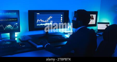 Homme Trader africain travaillant à l'intérieur du bureau la nuit portant casque de réalité virtuelle - concept de trading et de technologie - Soft focus sur sa tête Banque D'Images