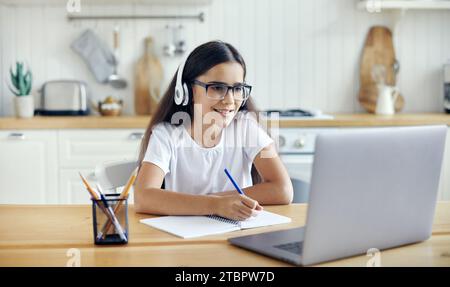 Pré-adolescent 12s fille dans des écouteurs et des lunettes assis à la table e-learns, écouter le cours en ligne, leçon audio, acquérir de nouvelles connaissances, des compétences en utilisant Internet et Banque D'Images