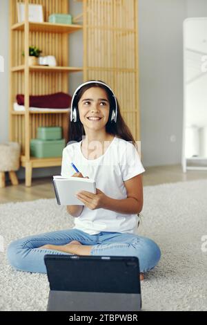 Jolie fille pré-adolescente assis sur le sol dans le salon confortable porter des écouteurs sans fil étudiant en ligne utiliser un appareil tablette numérique. Auto-éducation, jeune génération Banque D'Images