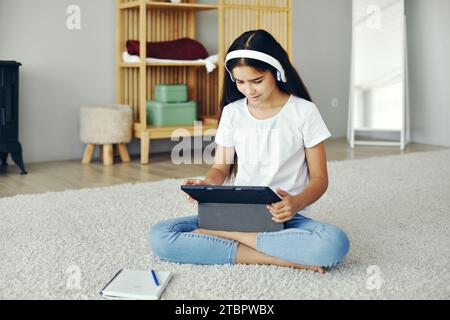 Jolie fille pré-adolescente assis sur le sol dans le salon confortable porter des écouteurs sans fil étudiant en ligne utiliser un appareil tablette numérique. Auto-éducation, jeune génération Banque D'Images