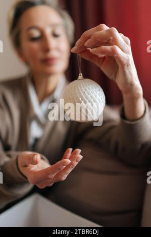 Cliché de mise au point sélective d'une femme d'âge moyen tenant et regardant une boule de Noël avec une expression heureuse. La femelle élégante décore l'arbre de Noël pour le nouvel an Banque D'Images