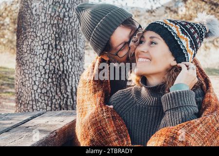 Couple heureux dans l'activité de loisirs romantique ensemble câliner au parc. L'automne aime les gens. Homme embrassant et liant femme mignonne assise sur un banc en bois avec des feuilles jaunes et des arbres en arrière-plan Banque D'Images