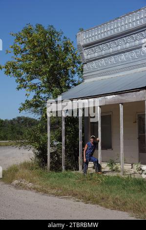 L'homme se penche contre le porche du vieux magasin de campagne sur la route de terre dans le Kansas. La façade du magasin est recouverte d'une boîte décorative. Banque D'Images