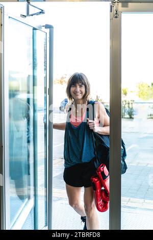 Photo verticale caucasienne mi-adulte femme portant des vêtements et de l'équipement de sport, entrant avec sourire par la porte à la salle de gym de boxe. Sport et recre Banque D'Images