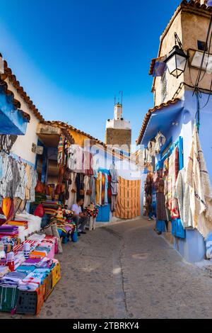 Chefchaouen, Maroc. 14 octobre 2022 - rue étroite avec boutiques de souvenirs et tissus typiques sur les façades bleues des maisons, avec le minaret o Banque D'Images