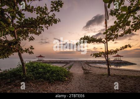 Lever du soleil sur la mer, vue de la plage de sable à l'horizon, dans la mer il y a des brise-lames avec de petits temples, eau calme avec de petites vagues et reflets sur la plage tropicale de Sanur, Bali, Indonésie Banque D'Images