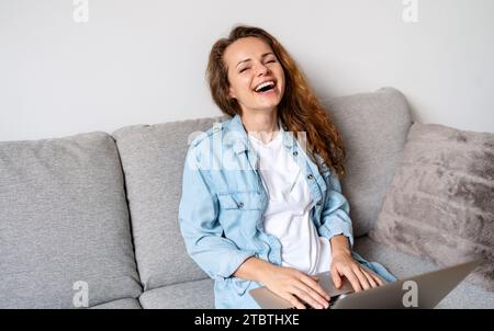 Femme riant alors qu'elle est assise sur le canapé à la maison et en utilisant son ordinateur portable. Banque D'Images