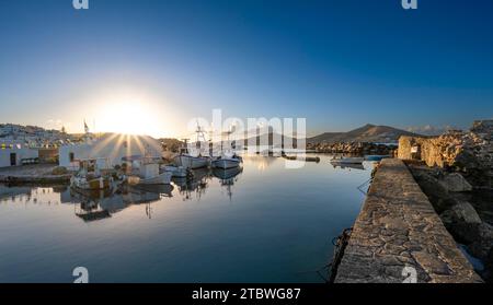 Bateaux de pêche dans le port de Naoussa au coucher du soleil, reflétés dans la mer, étoile du soleil, ruines du château vénitien sur la droite, maisons blanches des Cyclades Banque D'Images