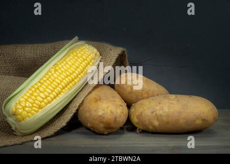 Matières, pommes de terre non pelées et les épis s'échappant du sac en toile de jute sur une vieille table en bois rustique, mur noir en arrière-plan, low angle view Banque D'Images