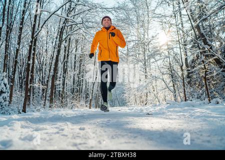 Homme de coureur de Trail souriant d'âge moyen vêtu d'une veste coupe-vent orange vif endurance course rapide forêt enneigée pittoresque pendant une journée ensoleillée gelée. S Banque D'Images