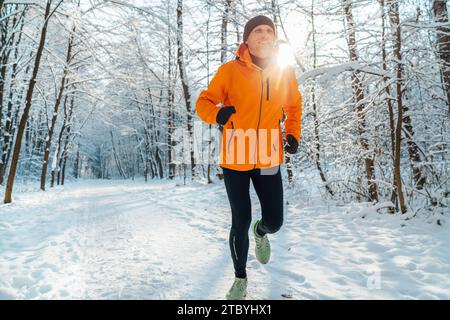 Homme de coureur de Trail souriant d'âge moyen vêtu d'une veste coupe-vent orange vif endurance course rapide forêt enneigée pittoresque pendant une journée ensoleillée gelée. S Banque D'Images