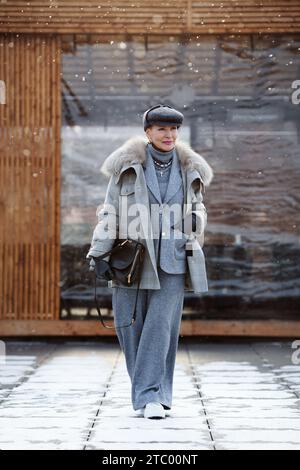 Femme mature élégante marche sur toute la longueur dans la rue en hiver, porte une tenue multicouche à la mode, manteau veste en laine grise, costume avec pantalon et col roulé Banque D'Images