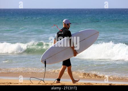 Homme portant planche de surf marchant vers la mer, El Cotillo, Fuerteventura, Îles Canaries, Espagne. Prise en novembre 2023. cym Banque D'Images