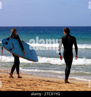 Homme et femme en combinaison avec planche de surf marchant vers la mer, El Cotillo, Fuerteventura, Îles Canaries, Espagne. Prise en novembre 2023 Banque D'Images