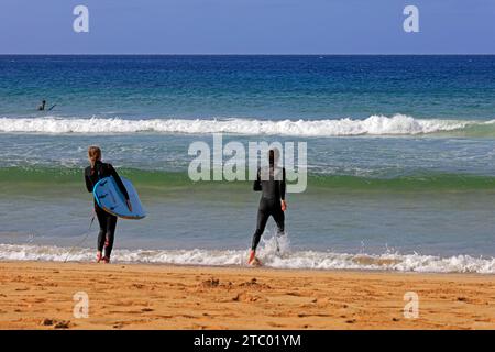 Homme et femme en combinaison avec planche de surf marchant vers la mer, El Cotillo, Fuerteventura, Îles Canaries, Espagne. Prise en novembre 2023 Banque D'Images