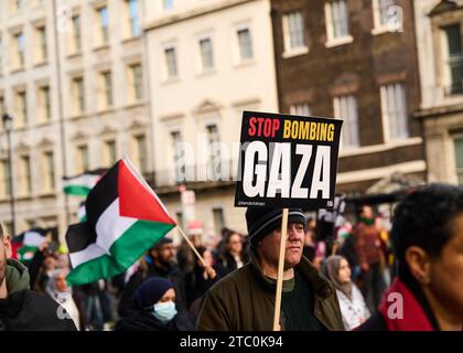 9 décembre 2023 - Londonuk : manifestant pro palestinien tenant une pancarte disant d'arrêter de bombarder gaza marchant à londres. drapeau palestinien en arrière-plan Banque D'Images