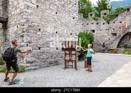 Un homme photographie ses fils alors que l'on prétend être prisonnier dans une réplique de pilori au milieu des ruines du château médiéval de Vezio à Perledo, Lombardie, Italie. Banque D'Images