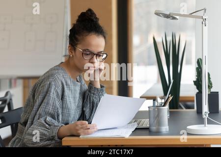 La travailleuse ethnique pensive lisait la paperasserie au bureau Banque D'Images