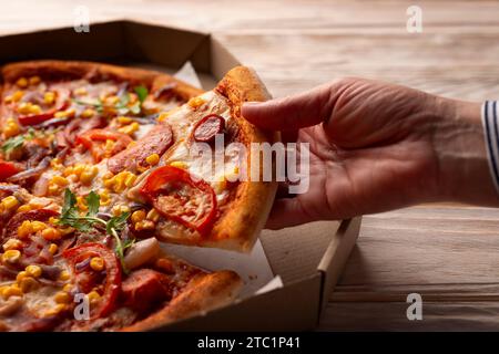 La main caucasienne humaine prend une tranche de grande pizza de la boîte de carton sur la table en bois blanc Banque D'Images