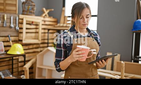 Jolie jeune femme charpentière blonde, café à la main, navigation touchpad au milieu de l'agitation de menuiserie dans l'atelier animé de menuiserie Banque D'Images