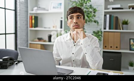 Concentré jeune homme hispanique immergé dans la pensée tout en tapant sur ordinateur portable, incarnant le succès au bureau. doutant mais confiant, cet ordinairement atlas Banque D'Images