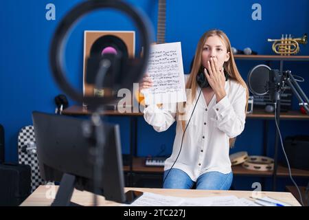 Jeune femme caucasienne faisant tutoriel de musique en ligne montrant la feuille de musique couvrant la bouche avec la main, choquée et effrayée pour erreur. expression surprise Banque D'Images