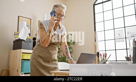 Confiante artiste chevronnée aux cheveux gris, cette artiste multitâche s'exprime sur son smartphone tout en utilisant son ordinateur portable dans son studio d'art inspirant. Banque D'Images