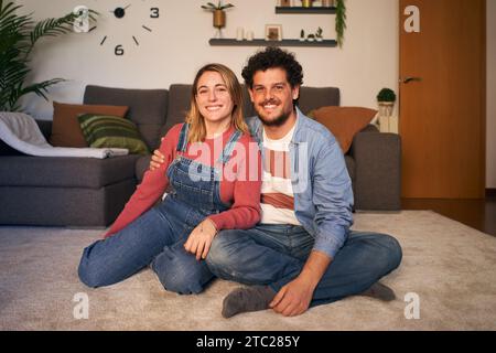 Deux caucasiens joyeux jeune adulte amoureux regardant une caméra souriante assise sur un tapis dans le salon. Banque D'Images