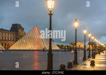 Pyramide du Louvre et palais en arrière-plan avec de vieux lampadaires alignés au premier plan illuminés en fin d'après-midi pluvieux. Banque D'Images