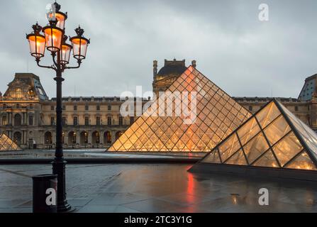 Musée du Louvre, Paris, France. Pyramide du Louvre et palais en arrière-plan avec une vieille lampe au premier plan illuminée en fin d'après-midi pluvieux. Banque D'Images