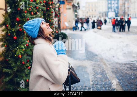 Heureuse jeune femme admire la chute de neige marchant sur la rue de la ville européenne décorée pour Noël portant des vêtements chauds. Espace Banque D'Images