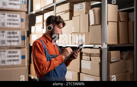 L'opérateur d'entrepôt masculin caucasien livrant des marchandises habillées de combinaisons bleues tient une tablette dans ses mains et prend des notes dessus. Le concept de Banque D'Images