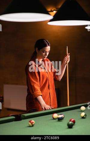 Une jeune femme jouant au billard se tient pensivement près de la table de billard avec une queue dans ses mains. Banque D'Images