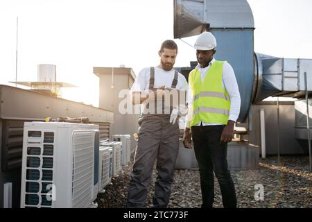 Superviseur afro-américain et technicien caucasien debout l'un à côté de l'autre et regardant l'écran de tablette moderne. Les ouvriers qualifiés de l'usine réintègrent Banque D'Images