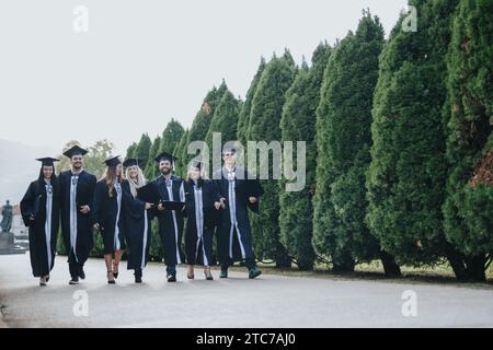 Les étudiants universitaires en robes de fin d'études célèbrent dans un parc ensoleillé, s'amusent et se félicitent mutuellement de leur réussite. Banque D'Images