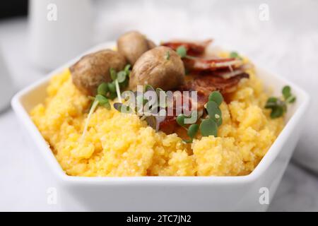 Semoule de maïs cuite avec bacon, champignons et microgreens dans un bol sur une table blanche, gros plan Banque D'Images